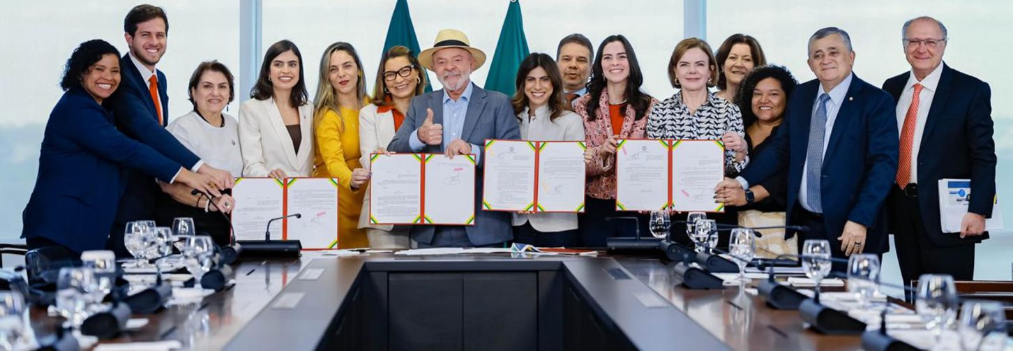 Diversas autoridades, com o presidente Lula ao centro, viradas para quem vê a foto, segurando o papel assinado da nova lei, em sala de reunião no Palácio do Planalto