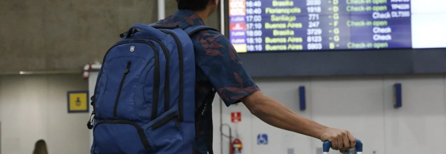 Passageiro com mochila nas costas e mala de rodinhas ao lado, em pé, observando painel de chegadas e partidas de voos em saguão de aeroporto