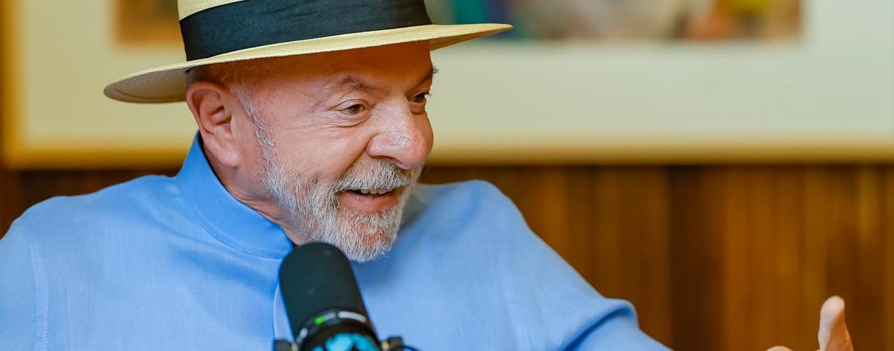 Presidente Lula de chapéu e camisa social azul, em close, sorridente, dando entrevista, microfone próximo e apoiado na mesa