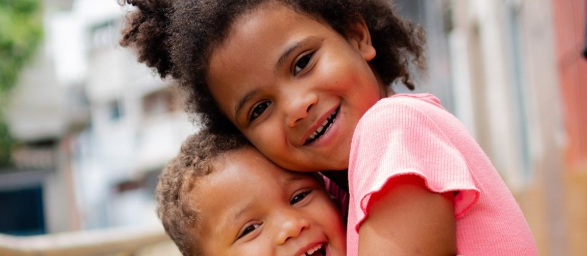 Foto de duas crianças pardas, sorrindo e se abraçando, um menino menor de camisa amarela e cabelo curto e uma menina maior de camisa rosa e cabelo encaracolado