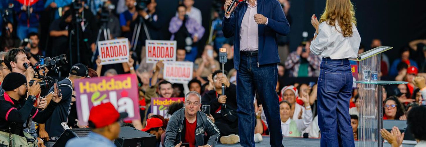 Fernando Haddad, de paletó e calça azul marinho, com camisa social rosa, caminhando em palanque, durante a convenção que oficializou o ex-ministro da Fazenda como candidato ao Governo de São Paulo, com pessoas ao redor e abaixo do patamar do palanque, com cartazes e de blusas vermelhas