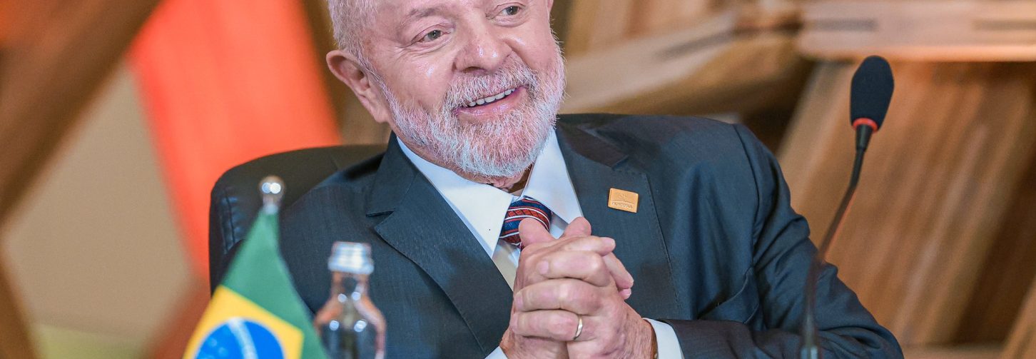 Presidente Lula, sentado, com uma mesa de madeira à frente e um pequeno suporte em cima da mesa com a bandeira do Brasil
