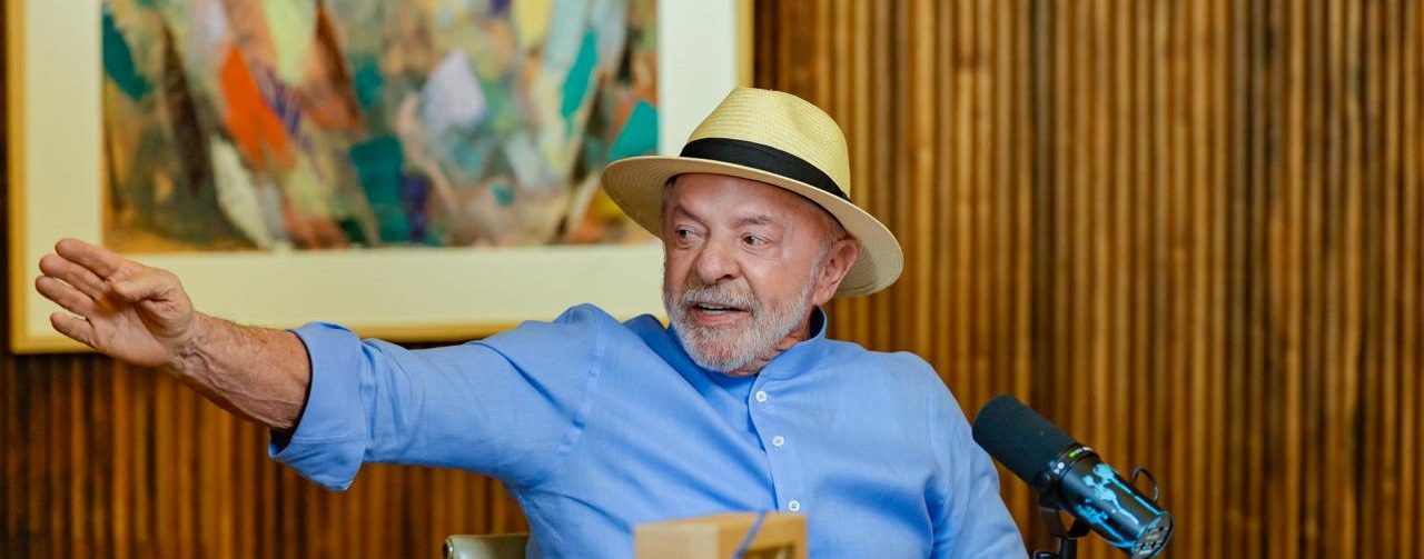 Presidente Lula de chapéu e camisa social azul, sentado dando entrevista