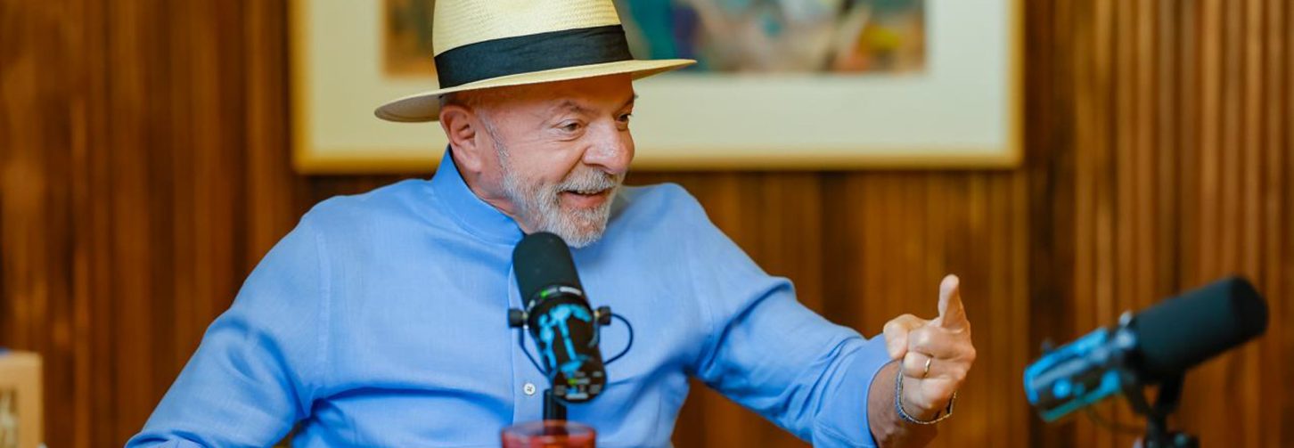 Presidente Lula de chapéu e camisa social azul atrás de mesa de madeira em entrevista a podcast no Palácio do Planalto