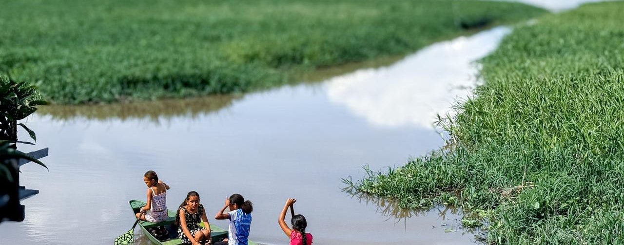 Quatro pessoas navegam em uma pequena canoa por um rio de águas barrentas. Uma delas conduz a embarcação com um remo. O curso d’água, cercado por extensa vegetação verde, faz uma curva e segue em direção ao horizonte.