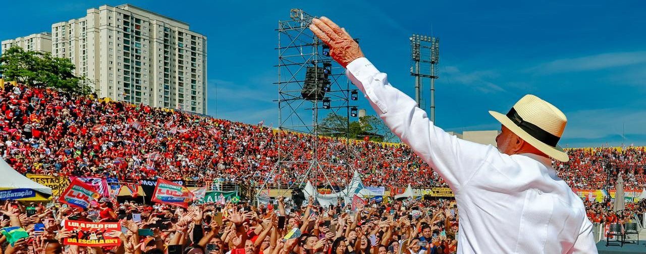 O presidente Lula em cima do palco, de costas para a foto, acena para multidão no estádio da Vila Euclides