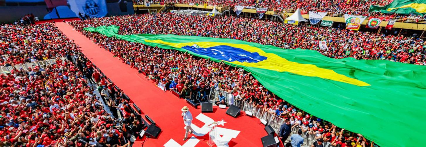 Vista aérea da Vila Euclides, com destaque para a passarela em que o presidente discursou, o público que enche o estádio e uma grande bandeira do Brasil estendida sobre parte do público