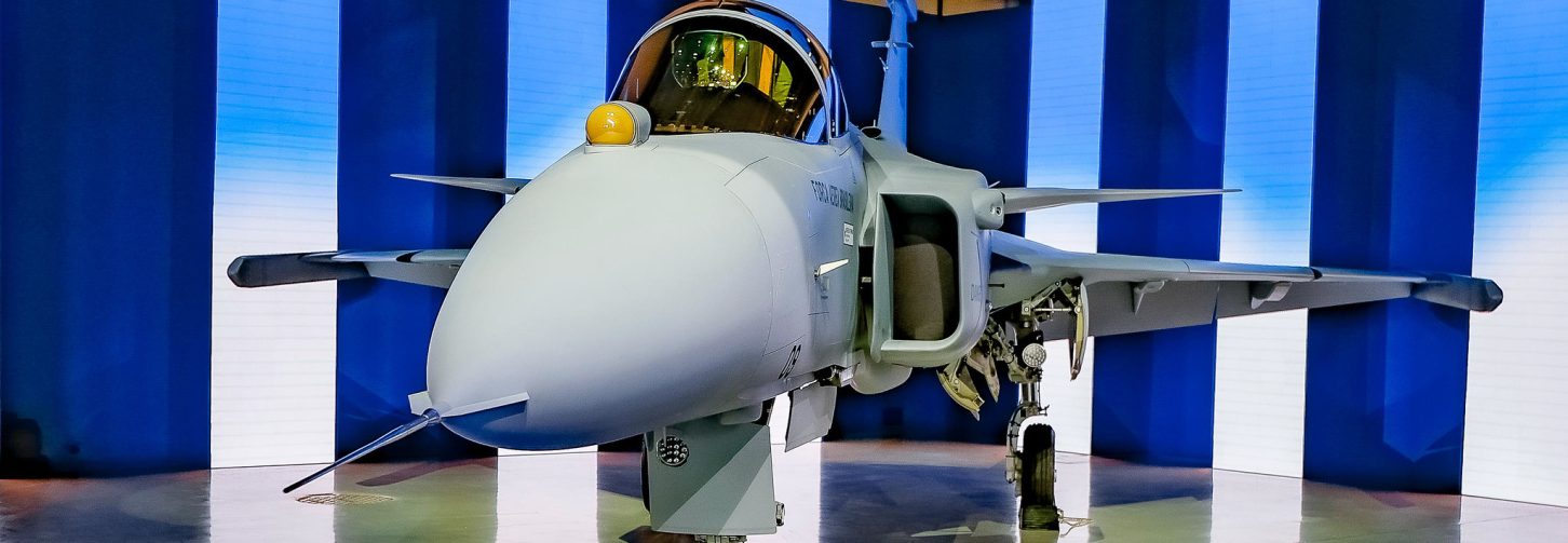 Em um espaço interno, um caça Gripen, da Força Aérea Brasileira, visto de frente, com o trem de pouso apoiado sobre o piso claro e brilhante. A aeronave tem asas curtas e cabine com cobertura transparente. Ao fundo, há painéis azuis e uma escada metálica ao lado do avião.