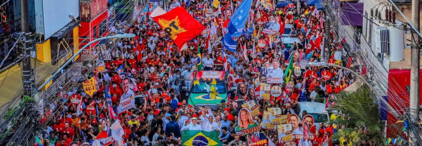 Imagem mostra uma grande mobilização de pessoas em torno do carro onde está o presidente Lula e integrantes da chapa majoritária na Bahia. Muitas bandeiras do Brasil, do PT, cartazes de candidatos muita gente nos prédios ao lado da rua assistindo a passagem do comboio
