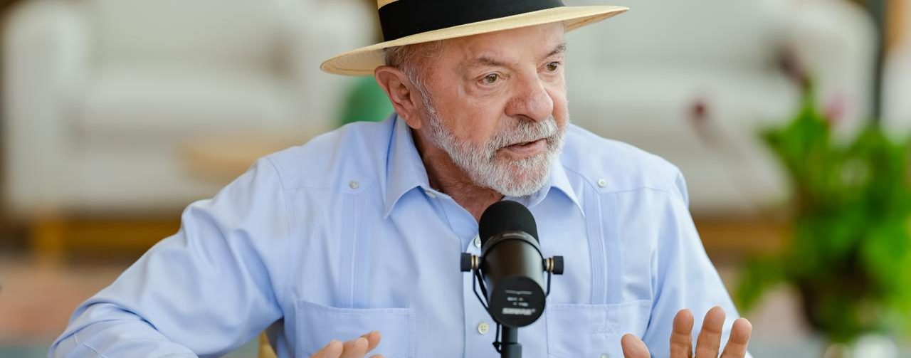 Presidente Lula, sentado em frente à microfone, veste camisa azul claro e chapéu Panamá