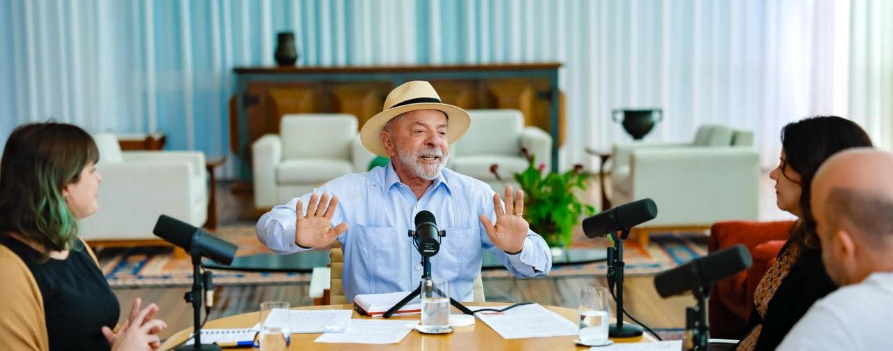 Lula sentado à uma mesa, vestindo camisa azul claro e chapéu Panamá, Ao seu lado esquerdo está uma mulher de cabelo castanho e blusa preta de mangas compridas e homem careca, de camiseta branca. Ao lado direito do presidente está outra mulher de cabelo castanho, vestindo uma blusa preta de mangas bege