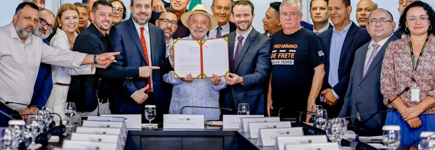 Lula, de camisa azul claro e chapéu, ao centro da imagem segurando a Lei do frete sancionada. do seu lado direito Guilherme Boulos, de terno azul escuro e gravata vermelha. Ao redor, lideranças e entidades, representantes do setor de transporte rodoviário de cargas
