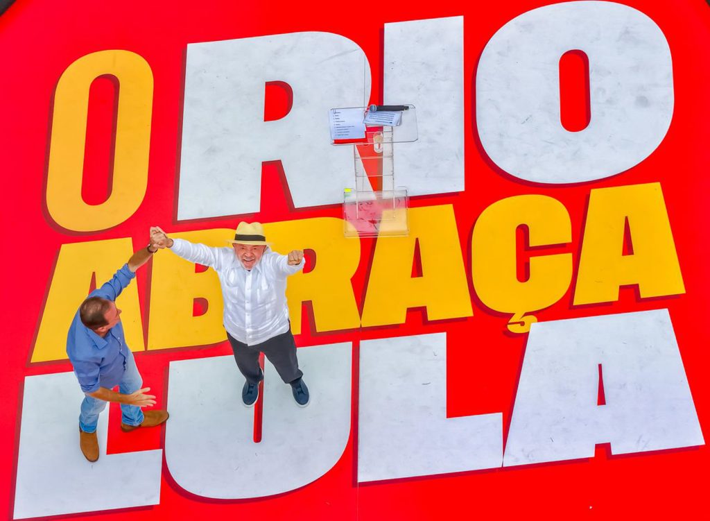 Vista de cima de Lula e Eduardo Paes sobre um palco vermelho. Lula usa camisa branca e chapéu claro, olha para o alto e ergue os braços; com uma das mãos, segura a mão do homem ao seu lado. No chão, em letras gigantes, está escrita a frase “O Rio abraça Lula”. Ao fundo, há um púlpito transparente.