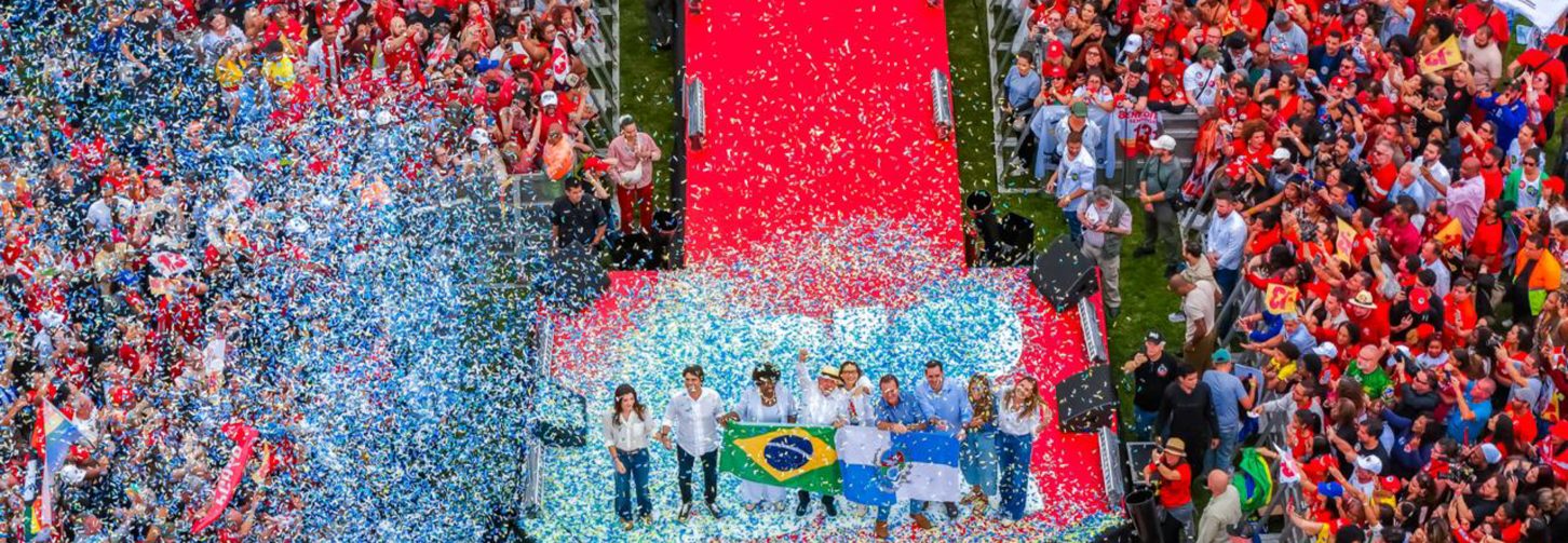 Vista aérea de uma multidão vestida predominantemente de vermelho, reunida ao redor de um palco e de uma passarela vermelha. No centro, pessoas seguram uma bandeira do Brasil enquanto uma chuva de papéis azuis, brancos e vermelhos cobre o espaço