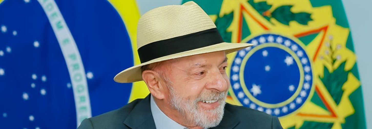 Presidente Lula, sentado atrás de uma mesa e com as bandeiras do Brasil e com o brasão ao fundo