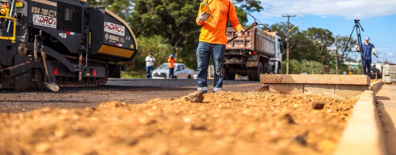 Em uma obra de pavimentação, um trabalhador caminha sobre o solo de terra ao lado de uma guia de concreto. Ele usa capacete amarelo, uniforme laranja e calça azul. Ao fundo, há máquinas de construção, outros trabalhadores, árvores e casas.