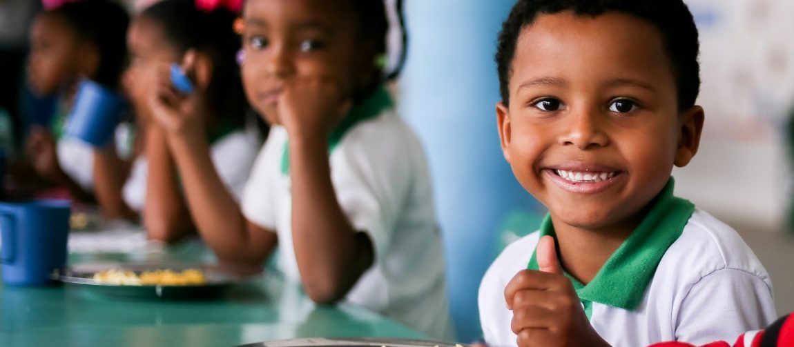 Em um refeitório escolar, um menino negro sorri para a câmera enquanto faz uma refeição. Ele veste uniforme branco com detalhes verdes e está sentado diante de uma bandeja com arroz, feijão e outros alimentos. Ao fundo, outras crianças também comem sentadas à mesa.