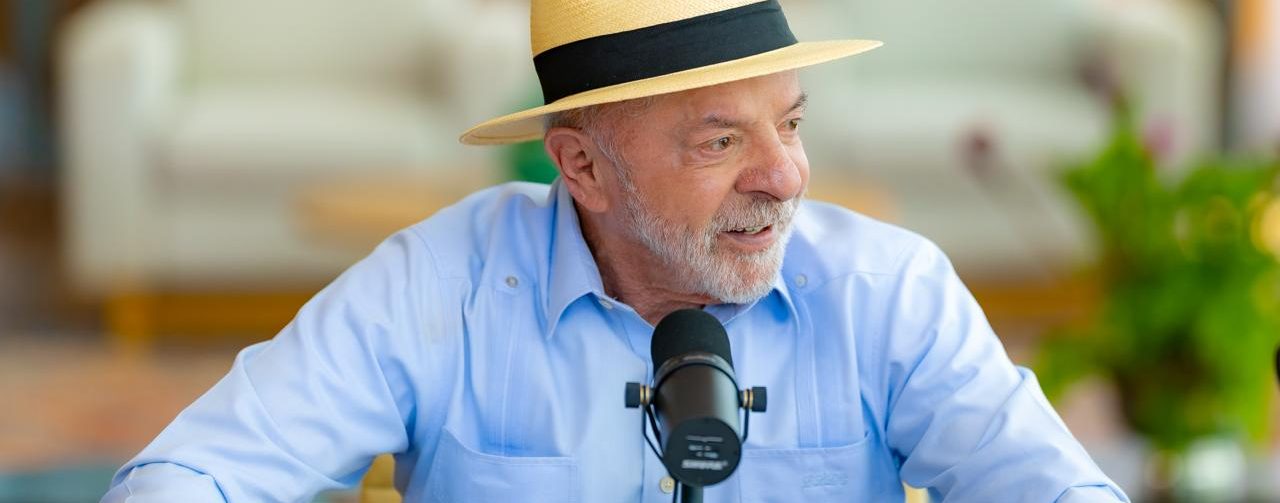 Lula, sentado em frente à microfone, vestindo camisa azul claro e chapéu Panamá