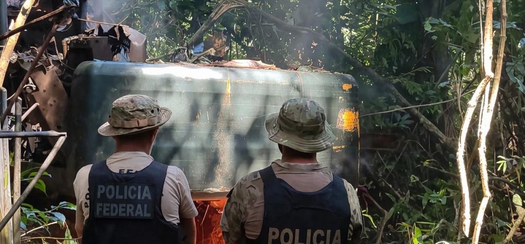 Em uma área de mata fechada, dois agentes vistos de costas observam uma estrutura metálica parcialmente escondida pela vegetação. Eles usam chapéus e coletes escuros com as inscrições “Polícia Federal” e “Polícia”. Ao redor, há troncos, galhos e folhagem densa.