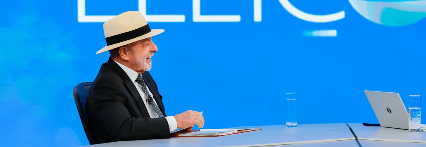 Lula está sentado diante de uma mesa durante uma entrevista. Ele usa terno escuro, camisa branca, gravata vermelha e chapéu de palha. À sua frente, há um microfone. Ao fundo, um painel azul exibe parte de uma inscrição em letras brancas.