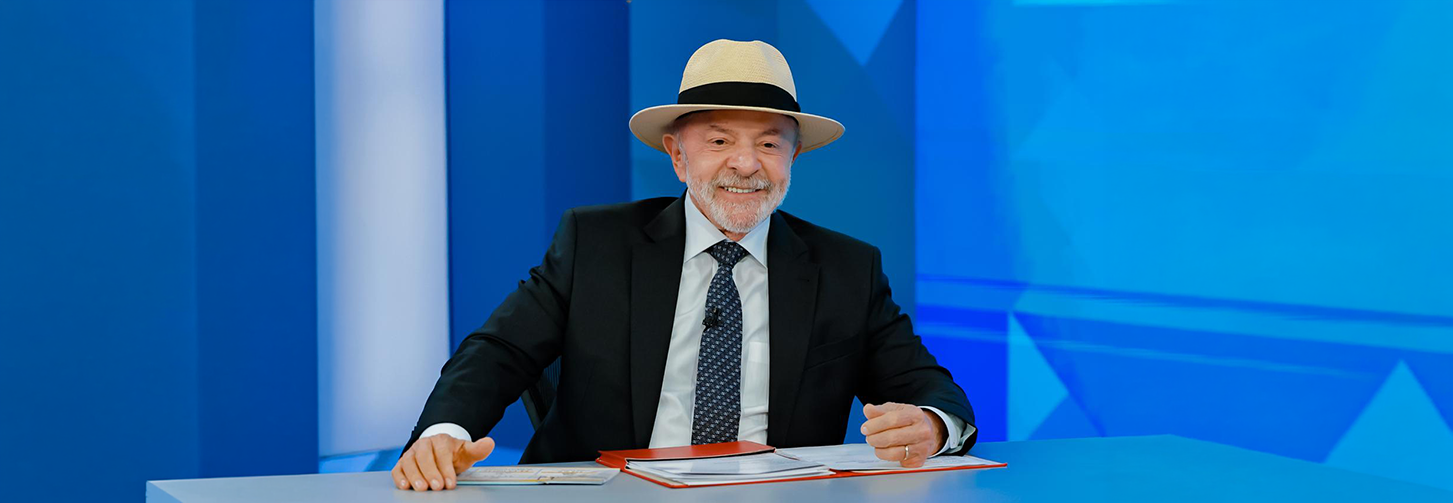Lula está sentado diante de uma mesa durante uma entrevista. Ele usa terno escuro, camisa branca, gravata vermelha e chapéu de palha. Ao fundo, um painel azul exibe parte de uma inscrição em letras brancas.