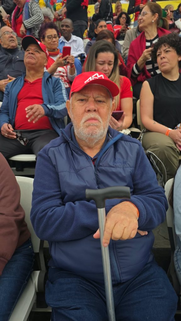 Vila Euclides- Adão fernandes (1) Um homem idoso, de barba grisalha, está sentado e olha para a câmera com expressão séria. Ele veste casaco azul e boné vermelho e apoia uma das mãos sobre uma bengala. Ao fundo, várias pessoas acompanham o evento, algumas vestidas de vermelho