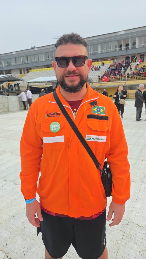 Um homem de cabelos curtos e barba posa para a câmera usando óculos escuros e uniforme laranja da Petrobras, com distintivos do SindiPetro e a bandeira do Brasil. Uma bolsa preta atravessa seu peito. Ao fundo, há arquibancadas com pessoas e um edifício de fachada cinza