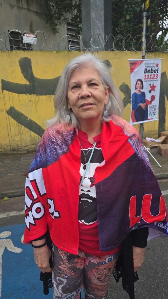 Uma mulher idosa, de cabelos grisalhos, posa para a câmera em uma rua. Ela veste uma camiseta vermelha e usa sobre os ombros uma bandeira em tons de vermelho, roxo e branco, com o nome “Lula” parcialmente visível. Ao fundo, há um muro amarelo e um cartaz de campanha.