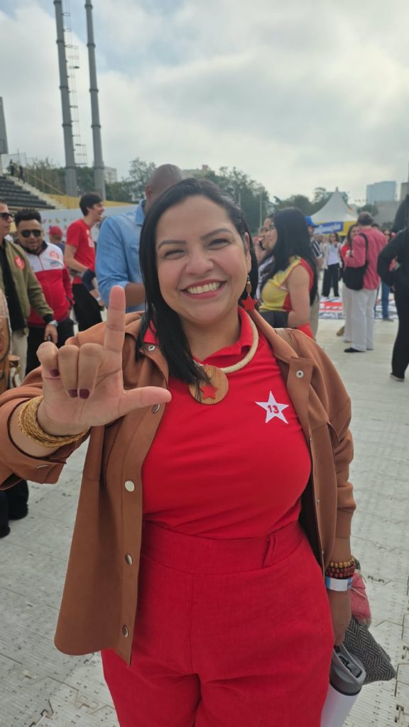 Uma mulher indígena de cabelos pretos sorri para a câmera e faz a letra L com a mão direita. Ela veste roupa vermelha com uma estrela branca e o número 13, além de uma jaqueta marrom e colares. Ao fundo, várias pessoas participam de um evento ao ar livre.