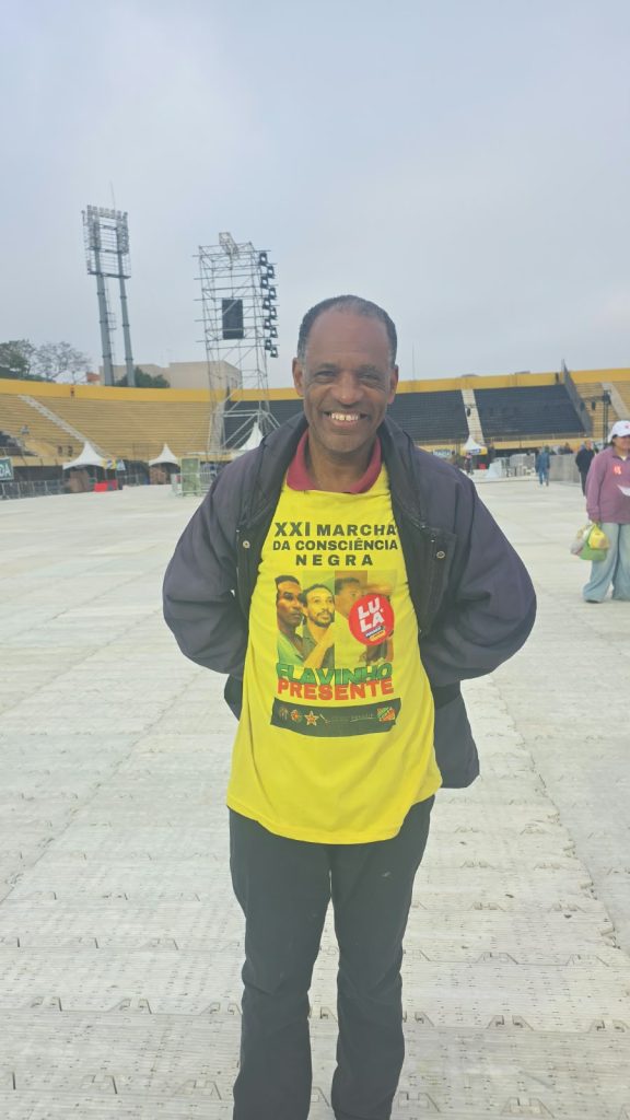 Um homem negro sorri para a câmera, com as mãos atrás do corpo. Ele veste casaco escuro sobre uma camiseta amarela com a inscrição “XXI Marcha da Consciência Negra”.  Ao fundo, há arquibancadas amarelas, estruturas de palco e algumas pessoas espalhadas pela arena.