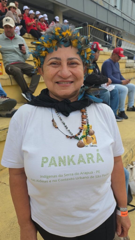 Vila Euclides- Delma Pancará Uma mulher indígena sorri para a câmera. Ela usa um cocar de penas azuis e amarelas, colares artesanais, camiseta branca com a palavra “Pankará” e um tecido preto amarrado sobre os ombros. Ao fundo, pessoas com roupas e bonés vermelhos ocupam uma arquibancada amarela.