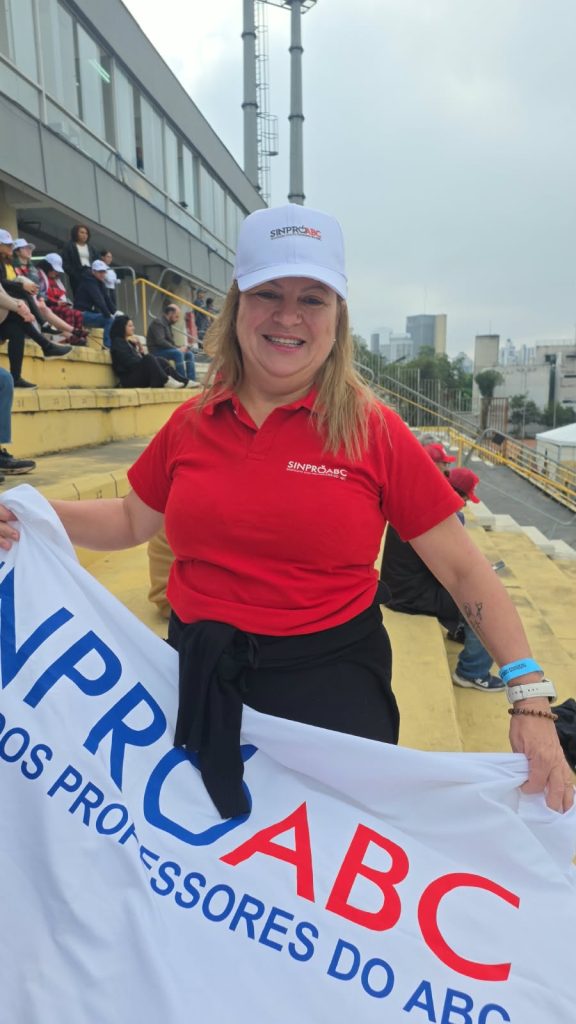 Vila Euclides- Edilene Uma mulher loira sorri para a câmera enquanto segura uma bandeira branca com as inscrições “Sinpro ABC” e “Sindicato dos Professores do ABC”. Ela veste camiseta vermelha e boné branco com o nome da entidade. Ao fundo, pessoas ocupam a arquibancada amarela de um espaço aberto.