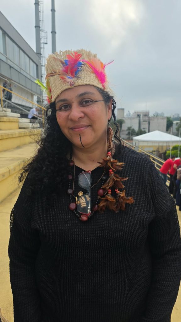 Vila Euclides – Elaine Pancará Uma mulher indígena de cabelos longos e cacheados olha para a câmera com um leve sorriso. Ela usa um adorno de palha com penas coloridas, colares artesanais e roupa preta. Seu rosto tem pinturas discretas nas bochechas e no queixo. Ao fundo, há arquibancadas amarelas, algumas pessoas e edifícios sob o céu nublado.