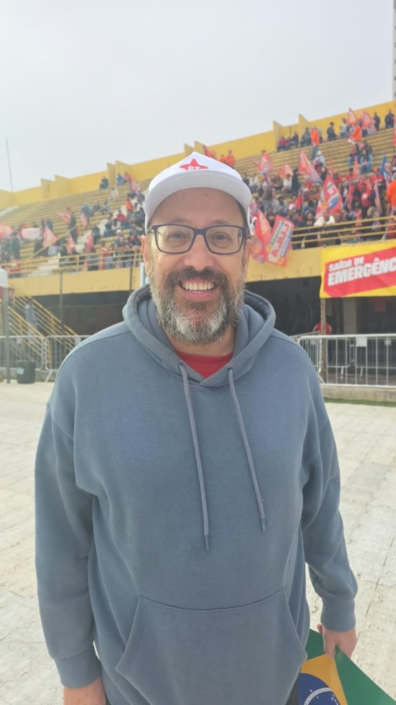 Vila Euclides – Fanley Bertoti Um homem de barba grisalha sorri para a câmera. Ele usa óculos, boné branco e moletom cinza. Ao fundo, uma multidão ocupa arquibancadas amarelas e segura bandeiras predominantemente vermelhas.