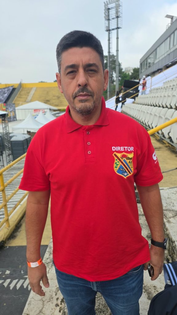 Vila Euclides- Haroldo(1) Um homem de cabelos curtos, bigode e barba rala olha para a câmera com expressão séria. Ele veste camisa polo vermelha com a palavra “Diretor” e um brasão bordado, além de calça jeans. Ao fundo, há arquibancadas brancas e amarelas e estruturas de um evento ao ar livre.