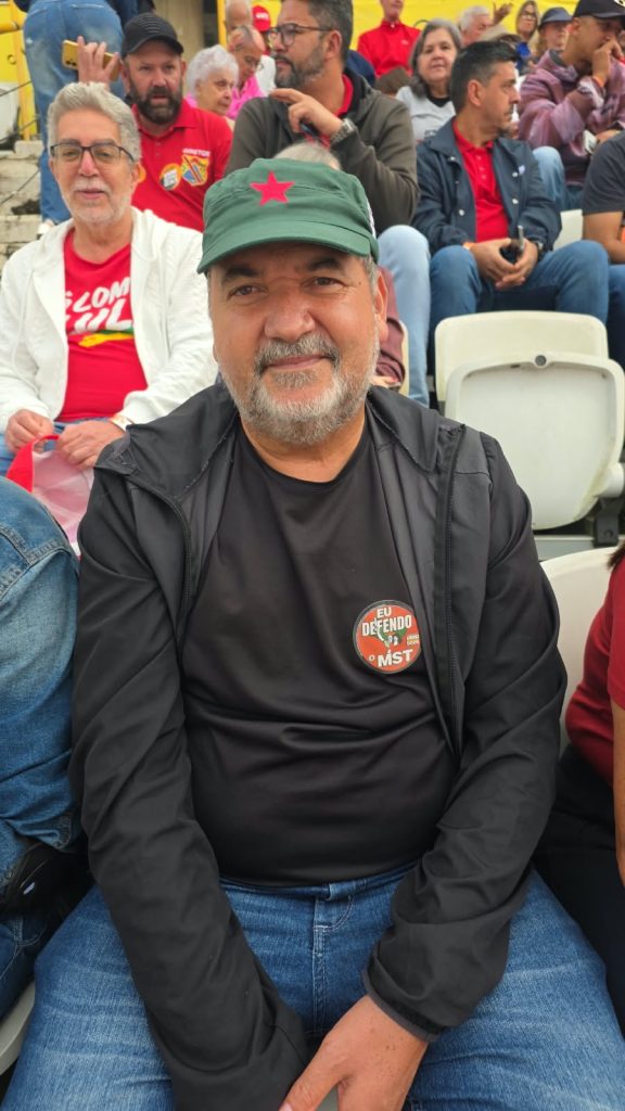 Um homem de barba grisalha sorri discretamente para a câmera, sentado em uma arquibancada. Ele veste roupas pretas e boné verde com uma estrela vermelha. Na camiseta, há um broche com a frase “Eu defendo o MST”. Ao fundo, outras pessoas acompanham o evento.