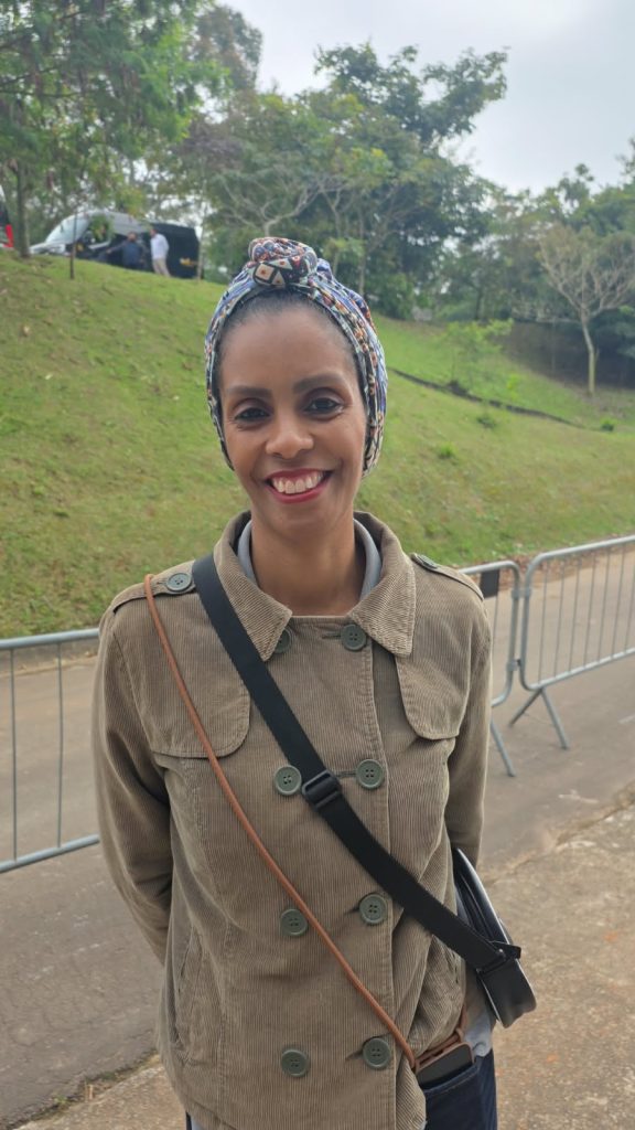 Uma mulher negra sorri para a câmera com as mãos atrás do corpo. Ela usa um lenço estampado envolvendo os cabelos, casaco marrom e duas bolsas atravessadas sobre o peito. Ao fundo, há grades metálicas, um gramado inclinado e árvores.
