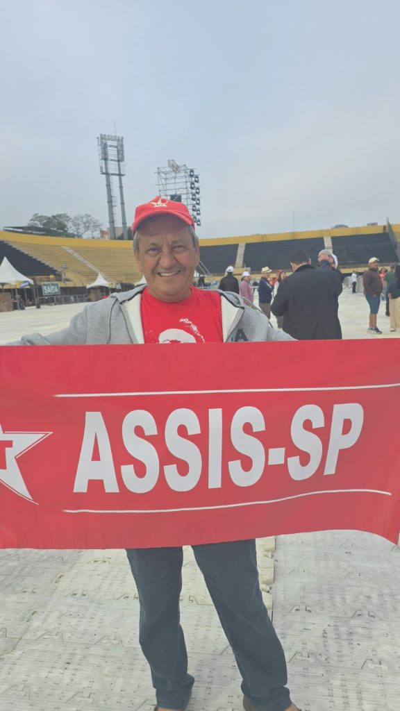 Vila Euclides- Luiz Carlos de Oliveira Um homem idoso sorri para a câmera enquanto segura uma faixa vermelha com a inscrição “PT Assis-SP” em letras brancas e o símbolo de uma estrela. Ele usa camiseta vermelha, casaco cinza e boné vermelho. Ao fundo, há pessoas espalhadas por uma arena com arquibancadas amarelas.
