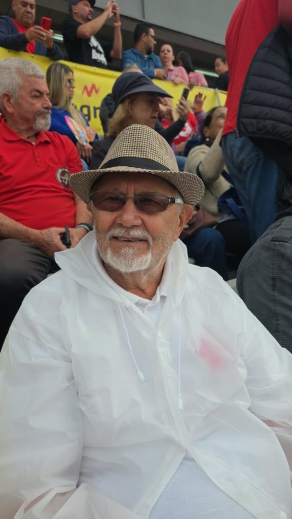 Um homem idoso, de barba e cabelos brancos, está sentado em uma arquibancada. Ele usa chapéu de palha, óculos escuros e capa de chuva transparente sobre uma camisa clara. Ao fundo, outras pessoas conversam, aplaudem e acompanham o evento.