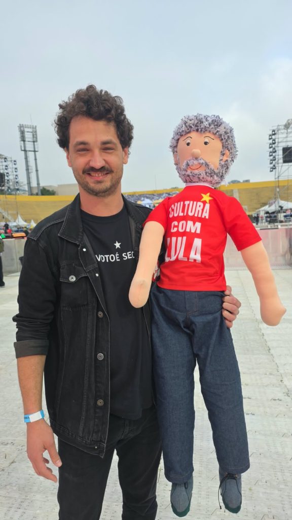 Vila Euclides – Michel Urânia Um homem de cabelos cacheados e bigode sorri para a câmera enquanto segura um boneco de pano que representa Lula. O boneco tem barba e cabelos grisalhos, veste calça azul e camiseta vermelha com a frase “Cultura com Lula”. Ao fundo, há uma arena com arquibancadas amarelas e estruturas de palco.