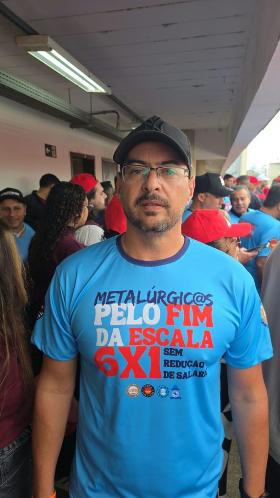 Vila Euclides- Rafael Loyola(1) Um homem de barba curta olha para a câmera. Ele usa óculos, boné preto e camiseta azul com a frase “Metalúrgicos pelo fim da escala 6x1, sem redução de salário”. Ao fundo, várias pessoas conversam em um corredor coberto.