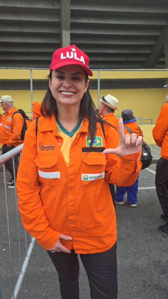Vila Euclides – Sandra Uma mulher de cabelos escuros sorri para a câmera. Ela usa uniforme laranja da Petrobras, com faixas refletivas e a bandeira do Brasil, e boné vermelho com a palavra “Lula”. Com a mão direita, faz a letra L. Ao fundo, outras pessoas também vestem uniformes laranja.