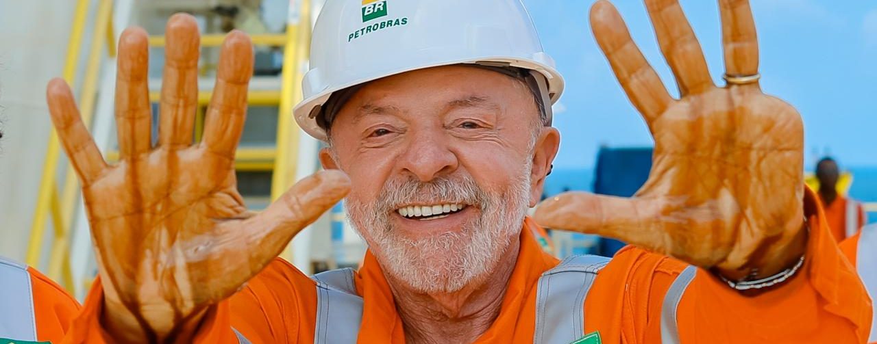 Presidente Lula com capacete branco com a marca da Petrobras e macacão laranja da empresa, sorrindo e mostrando as mãos sujas de petróleo