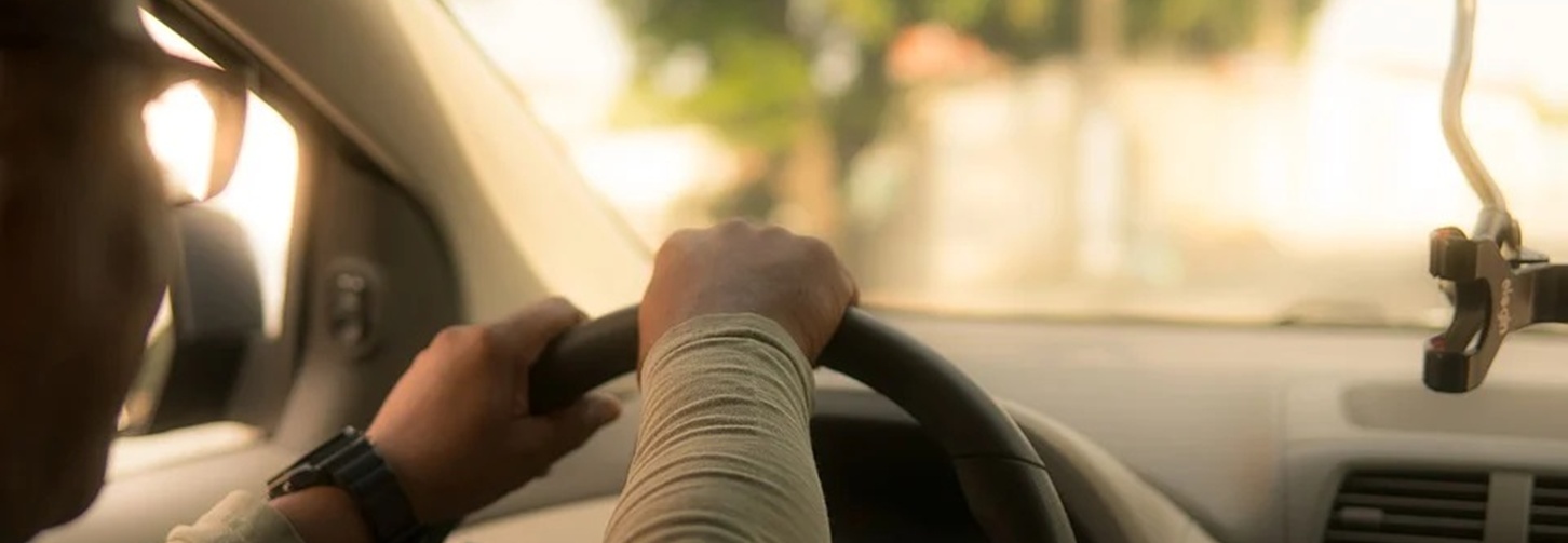 Vista do interior de um carro, por trás do motorista. Ele mantém as duas mãos ao volante enquanto dirige. À frente, a rua aparece desfocada e iluminada pela luz do sol.