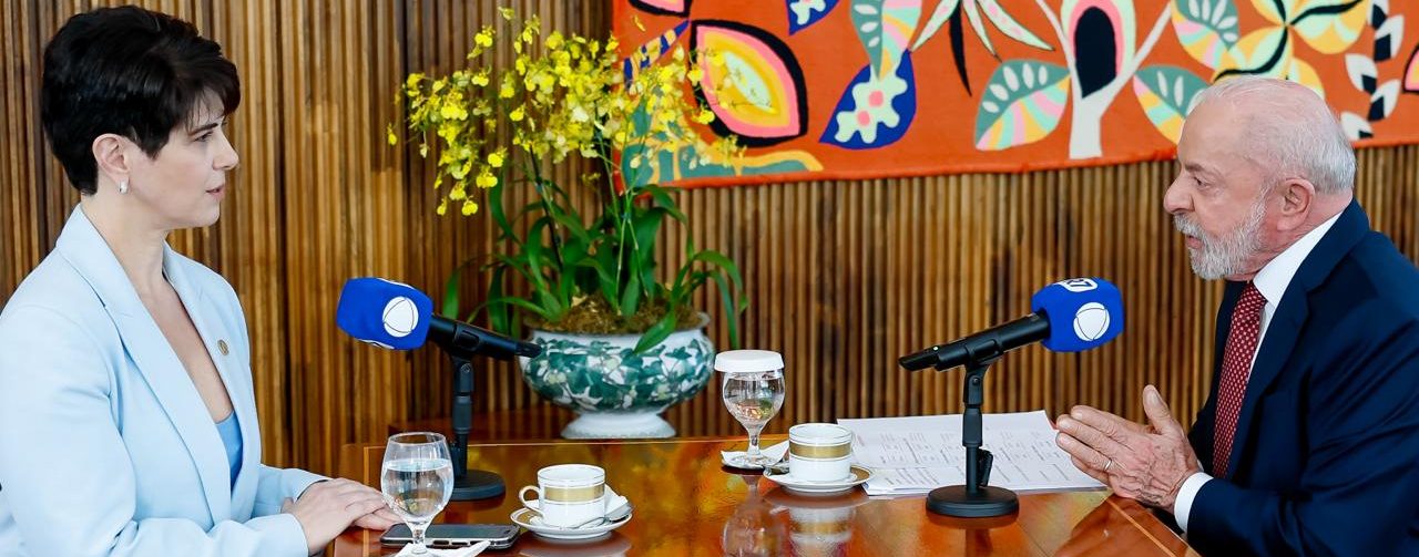Lula e a jornalista Mariana Godoy estão sentados frente a frente em uma mesa de madeira durante uma entrevista. Sobre a mesa há microfones, copos, papéis e um arranjo de flores. Ao fundo, um grande painel colorido decora a sala, que tem paredes e piso de madeira