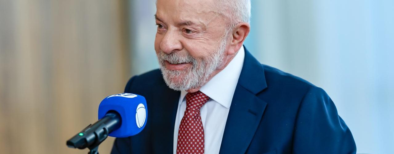 Lula está sentado à mesa durante uma entrevista. Ele veste terno azul, camisa branca e gravata vermelha, fala diante de um microfone e gesticula com as mãos. Ao fundo, há painéis decorativos coloridos.