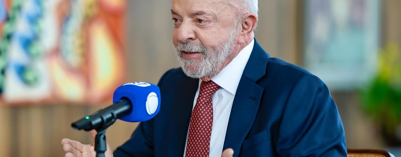 Lula está sentado à mesa durante uma entrevista. Ele veste terno azul, camisa branca e gravata vermelha, fala diante de um microfone e gesticula com as mãos. Ao fundo, há painéis decorativos coloridos.