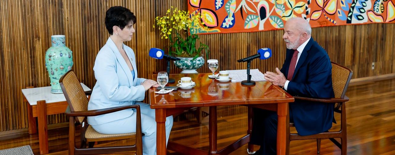 Lula e a jornalista Mariana Godoy estão sentados frente a frente em uma mesa de madeira durante uma entrevista. Sobre a mesa há microfones, copos, papéis e um arranjo de flores. Ao fundo, um grande painel colorido decora a sala, que tem paredes e piso de madeira.