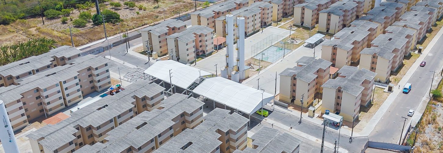 Vista aérea de um grande conjunto habitacional em construção. Diversos blocos de prédios, distribuídos em fileiras, estão em diferentes etapas da obra. Ao redor, há ruas, terrenos e áreas de vegetação.