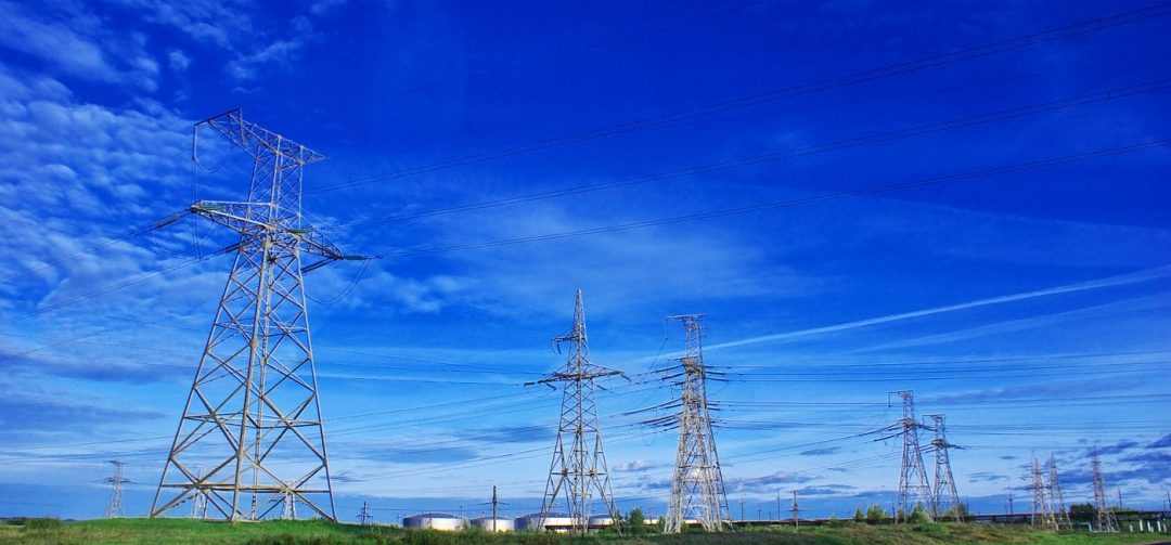 Torres de transmissão de energia estão distribuídas por uma extensa área de vegetação, ligadas por cabos elétricos. Em primeiro plano, uma estrada asfaltada atravessa a paisagem. Ao fundo, o céu azul tem nuvens finas e alongadas.